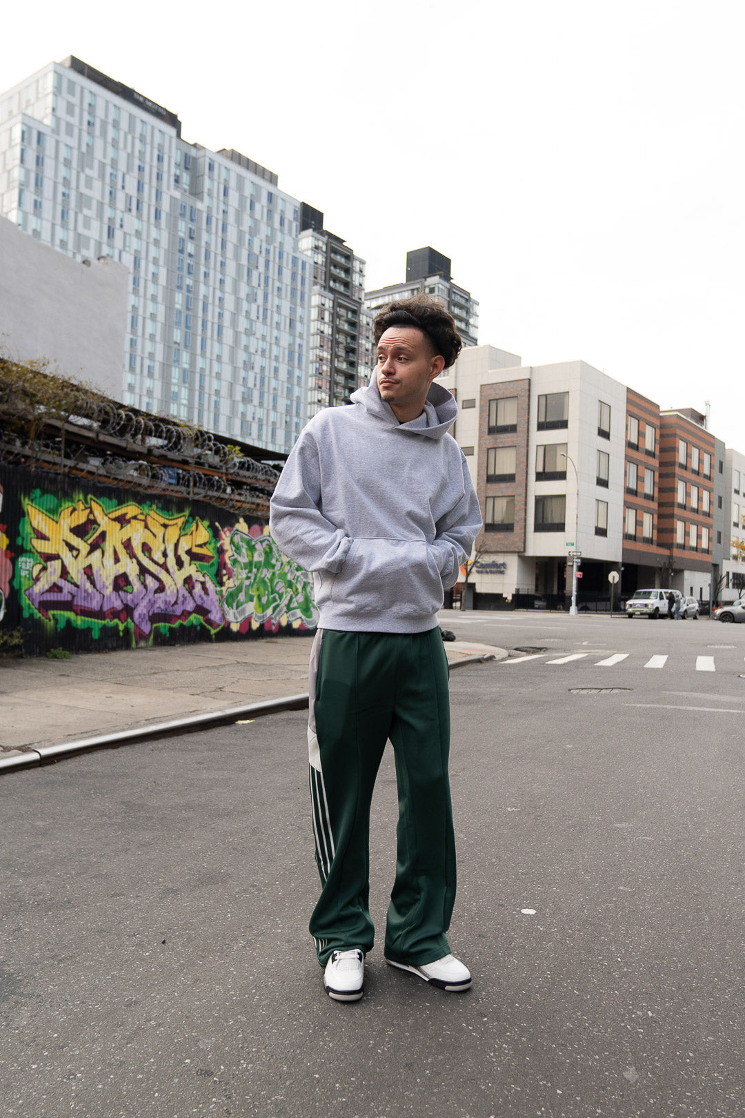 Person wearing a gray hoodie and green tracksuit standing on an urban street with graffiti.